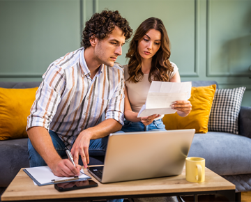 couple looking at paper and laptop while writing and sitting on the couch